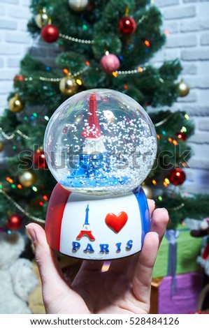 Snow ball in a man's hand on the background of the Christmas tree. Snow globe and Eiffel Tower inside. French decor.