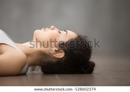 Side view portrait of beautiful young woman working out against grey wall, resting after doing yoga exercises, lying in Shavasana (Corpse or Dead Body Posture), relaxing. Face close up Royalty-Free Stock Photo #528602374