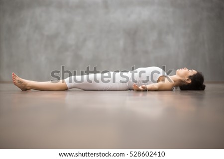 Side view portrait of beautiful young woman working out against grey wall, resting after doing yoga exercises, lying in Savasana (Corpse or Dead Body Posture), relaxing. Full length Royalty-Free Stock Photo #528602410