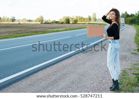 Young woman hitchhiker on the road is holding a blank cardboard sign 