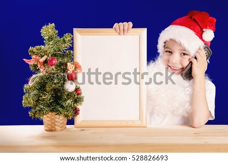 Young Santa Claus sitting at a wooden table holding a smart phone and an empty picture frame with a white background. He looks at the camera, smiling. Christmas tree on the table. Blue background.