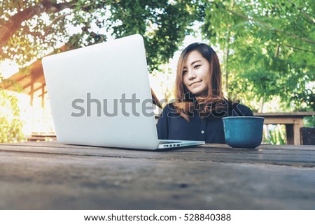 A beautiful asian woman using laptop with feeling happy and smiley face , sitting and looking in green nature outdoor background , hot coffee cup on wood table