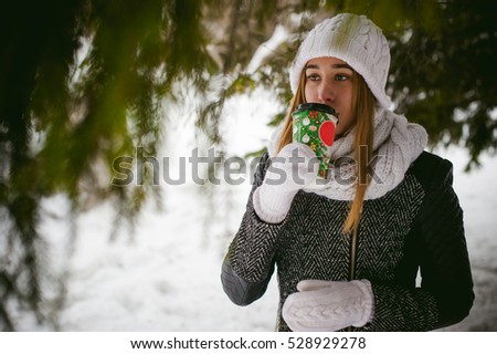 Portrait of cute woman in white scarf and hat knitted coat on outdoors background of snow and blurred fir branches in winter. girl holds a coffee cup in hand, christmas takeaway coffee