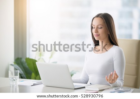 Young woman near the laptop, practicing meditation at the office desk, in front of laptop, online yoga classes, taking a break time for a minute, healing from paperwork and laptop radiation Royalty-Free Stock Photo #528930796