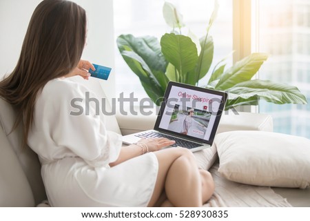 Young woman wearing dressing gown shopping online, sitting comfortably, using card for paying, wasting all spending, comparing prices, no legwork, safe and careful with websites to purchase. Rear view
