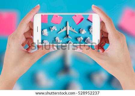 Woman hand using smart phone taking photo for preparation Traveling with airplane model flying among paper clouds and pink paper noted. Travel concepts, Ambient blurry background.