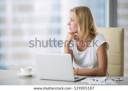 Young attractive woman at a modern office desk, working with laptop, looking at the window, thinking about a post, full-time blogger, seeking for inspiration, help to be productive, updating computer Royalty-Free Stock Photo #529005187