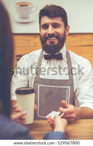 Young handsome barista with beard in shirt and bow tie giving a cup and taking a credit card while standing at the bar counter