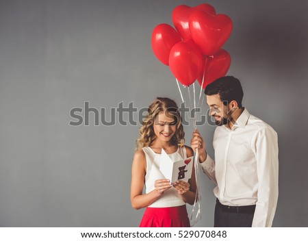 Handsome elegant guy is presenting a gift card and balloons to his beautiful girlfriend and smiling Royalty-Free Stock Photo #529070848