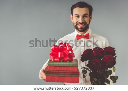 Handsome elegant guy in classic shirt with red bow tie is holding a gifts and roses, looking at camera and smiling, on gray background