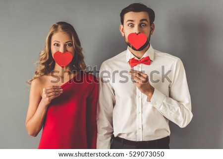 Beautiful elegant girl in red dress and guy in classic shirt and red bow tie are holding paper hearts, on gray background