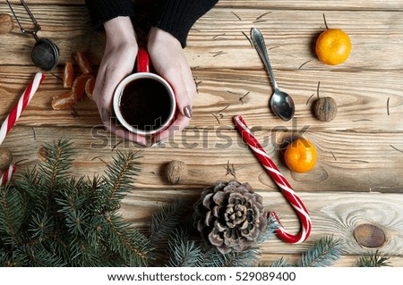 Women's hands with a neat manicure hold a cup on a background of a New Year's still-life. Woman in sweater heated a cup of tea near the tangerine and Christmas candies.
