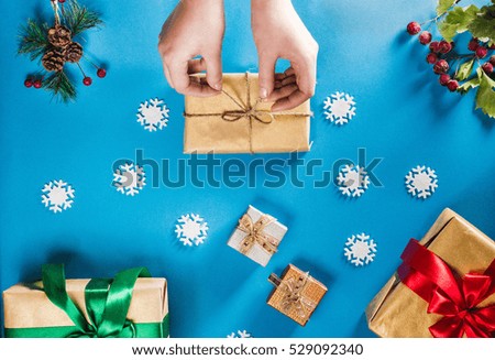 Concept of Christmas items on a bright blue background. Woman's hands wrapping gift. Flat lay decorations, snowflakes, berries and twigs with presents.