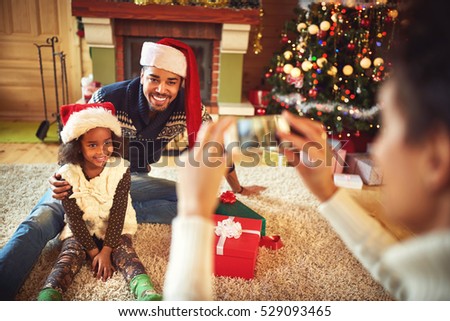 mother taking picture of smiling father and daughter in Santa hats at Christmas
