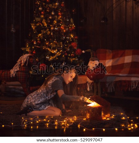Little girl on Christmas night opens a box with a gift. Magic, sparkles and light pour on her face. Background dark room with decorated spruce. Art warm picture. On the floor are little lights 