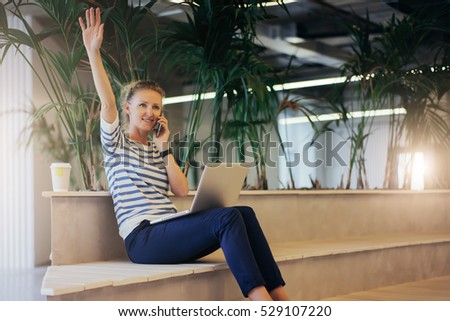 Young woman in striped T-shirt sitting in room with a modern interior and talking on cell phone.On lap of girl is a laptop.Girl welcomes friends raised hand.Girl uses gadget.In background houseplants.