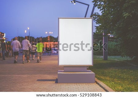 Summer evening. Front view. Blank vertical bilbor stands in city park. In the background, a walking group of people. Mock up. Advertising space.