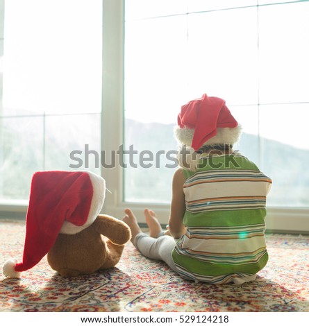 Christmas picture little boy in Santa hat with cute teddy bear sitting in front of window