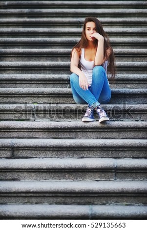 Stylish long-haired girl in a t-shirt and jeans sitting on stone stairs.