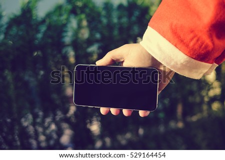 Santa Claus holding black modern model of mobile smartphone ready for Christmas time on green pine background