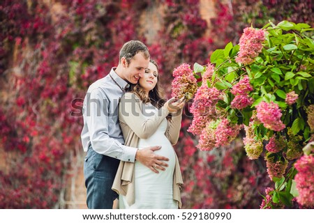 A pregnant young woman and her husband. A happy family standing at the red autumn hedge, smelling a flower hydrangea. pregnant woman relaxing in the park. Lovely prospective mother