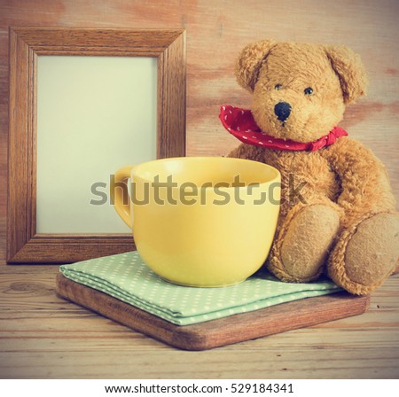 Yellow empty cup coffee on fabric with teddy bear sitting and frame picture. Wood background image vintage style. 