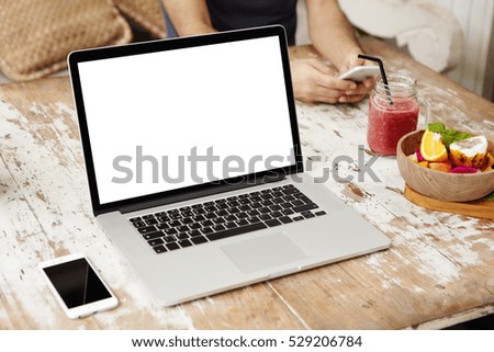 Generic modern laptop with blank copy space screen resting on wooden table with mobile phone, smoothie and fruits. Man using electronic gadget sitting on background. Selective focus. Mock up