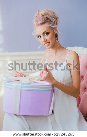 portrait of a luxurious blonde in a white dress sitting on a chair with a round gift box in hand on a lilac background