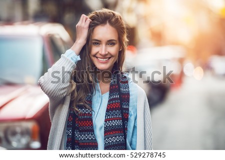 Happy young adult woman smiling with teeth smile outdoors and walking on city street at sunset time weating winter clothes and knitted scarf. Royalty-Free Stock Photo #529278745