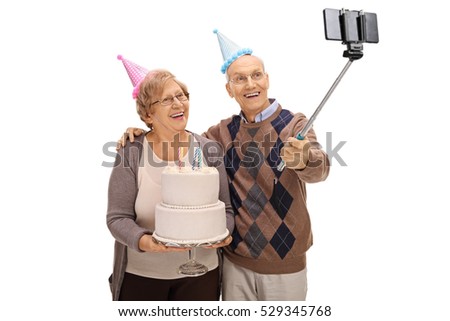 Joyful seniors with party hats and a birthday cake taking a selfie with a stick isolated on white background
