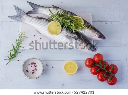 Fresh Mullet on a marble board over vintage wooden background with rosemary, lemon and tomatoes, seasoned with salt and rose pepper. Top View