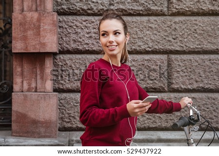 Picture of cheerful woman dressed in sweater walking with her bicycle in the city while standing near wall and listen to music by her phone. Look aside