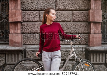 Picture of young woman dressed in sweater walking with her bicycle in the city while standing near wall. Look aside.