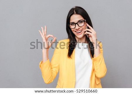 Photo of charming young woman wearing eyeglasses make Okay gesture while talking by her phone over grey background. Look at camera.