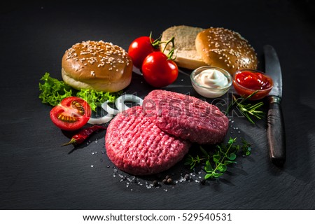 Raw beef patties, sesame buns with other ingredients for hamburgers on dark slate plate