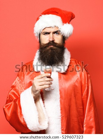 handsome bearded santa claus man with long beard on serious face holding glass of alcoholic shot in christmas or xmas sweater and new year hat on red studio background