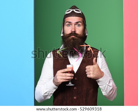handsome bearded pilot or aviator man with long beard and mustache on serious face holding glass of alcoholic shot in vintage suede leather waistcoat with hat and glasses on colorful studio background