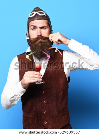 handsome bearded pilot or aviator man with long beard and mustache on funny face holding glass of alcoholic shot in vintage suede leather waistcoat with hat and glasses on blue studio background