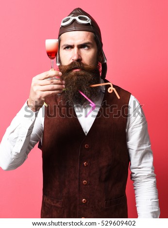 handsome bearded pilot or aviator man with long beard and mustache on serious face holding glass of alcoholic shot in vintage suede leather waistcoat with hat and glasses on red studio background