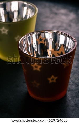 Vertical photo of two color semi-transparent cups with shiny surface from inside. Transparent stars are on side. Single small candle is placed in both and light goes through. Background is black plate