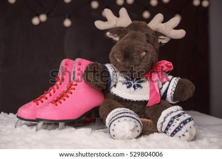 Winter holidays decoration. Pink skates for figure skating and toy elk or deer on the snow over dark background.