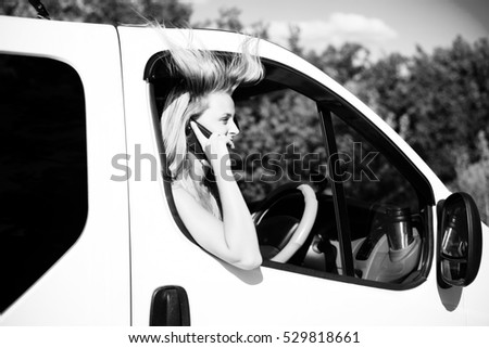 Black and white picture of emotional blond girl speaking phone and looking from car window. Young woman excited while talking with someone on sunny countryside background.