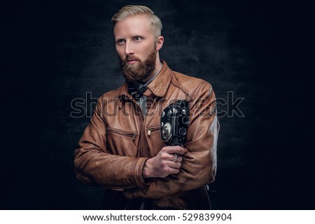 Portrait of blond bearded male dressed in brown leather jacket holds vintage 8mm video camera.