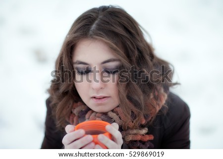 Beautiful young brunette girl looking down, holding a mug of tea in winter outdoors in the snow
