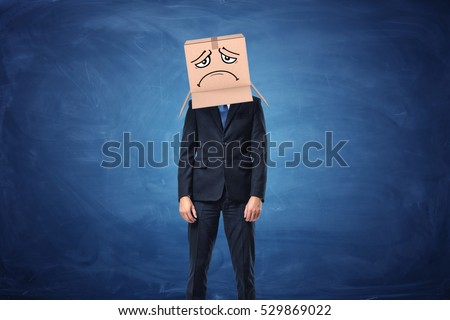 Businessman wearing cardboard box with drawn sad face on his head near the blue blackboard. Anonymous person. Packing and crating. Issues of small business. Royalty-Free Stock Photo #529869022