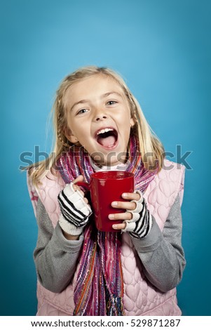 Young cheerful girl wearing winter clothing and drinking hot chocolate shot in the studio isolated on blue background.