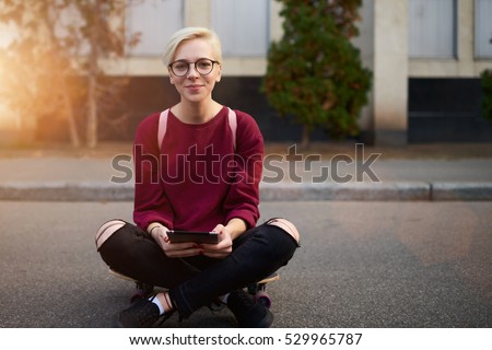 Portrait of young female skater having rest after engage sports activities in urban setting. Hipster girl holding touch pad with made photos of her ride. Copy space area for advertising text message