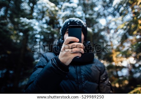 Man takes a picture on mobile phone in the snow-covered pine forest on a cold winter day