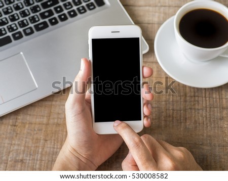 Close up business man's hand holds white smart phone with black isolated screen and points on the screen over background of laptop, coffee on wooden background