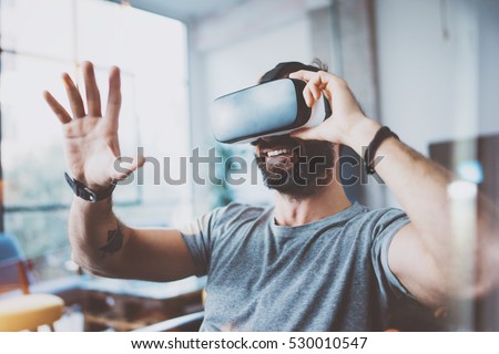 Young bearded man wearing virtual reality glasses in modern interior design coworking studio. Smartphone using with VR goggles headset. Horizontal,flares effect, blurred background Royalty-Free Stock Photo #530010547